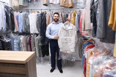 Photo of Dry-cleaning service. Happy worker holding hanger with jacket in plastic bag near other clothes indoors