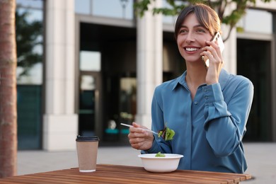 Happy businesswoman with plastic bowl of salad talking on smartphone during lunch at wooden table outdoors
