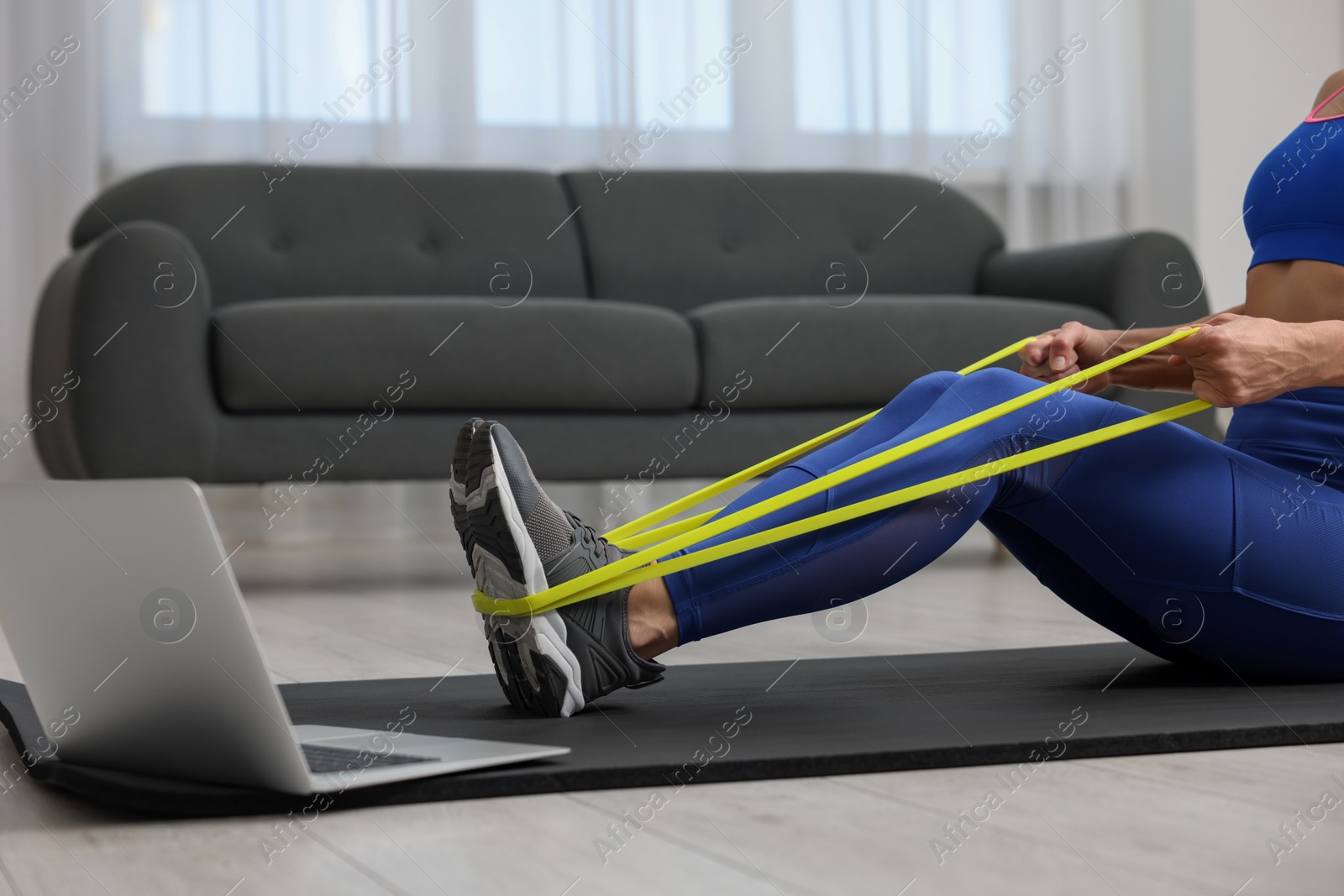 Photo of Woman doing exercise with fitness elastic band near laptop on mat at home, closeup