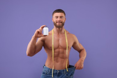 Photo of Athletic young man with measuring tape and bottle of supplements wearing big jeans on purple background. Weight loss