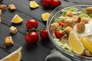 Photo of Bowl of delicious salad with Chinese cabbage, lemon, tomatoes and bread croutons on black wooden table, closeup