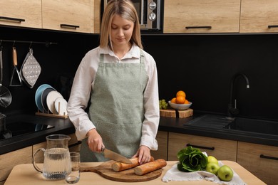 Young woman in clean apron cutting baguette on wooden table at kitchen
