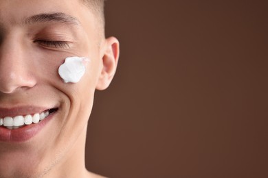 Handsome man with moisturizing cream on his face against brown background, closeup. Space for text