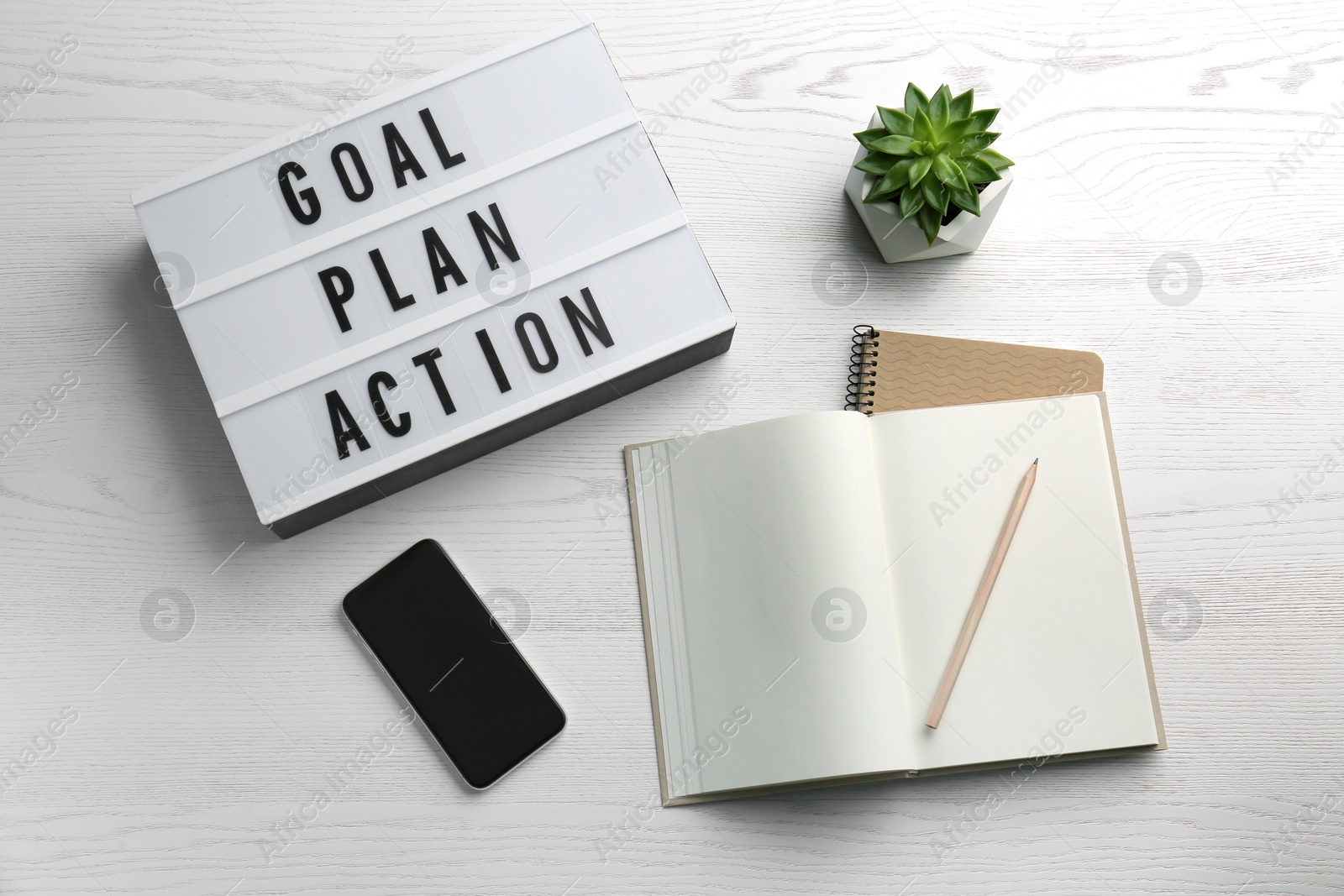 Photo of Light box with text Goal, Plan, Action near notebooks and smartphone on white wooden table, flat lay