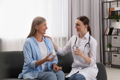 Photo of Young healthcare worker giving glass of water to senior woman with pills indoors