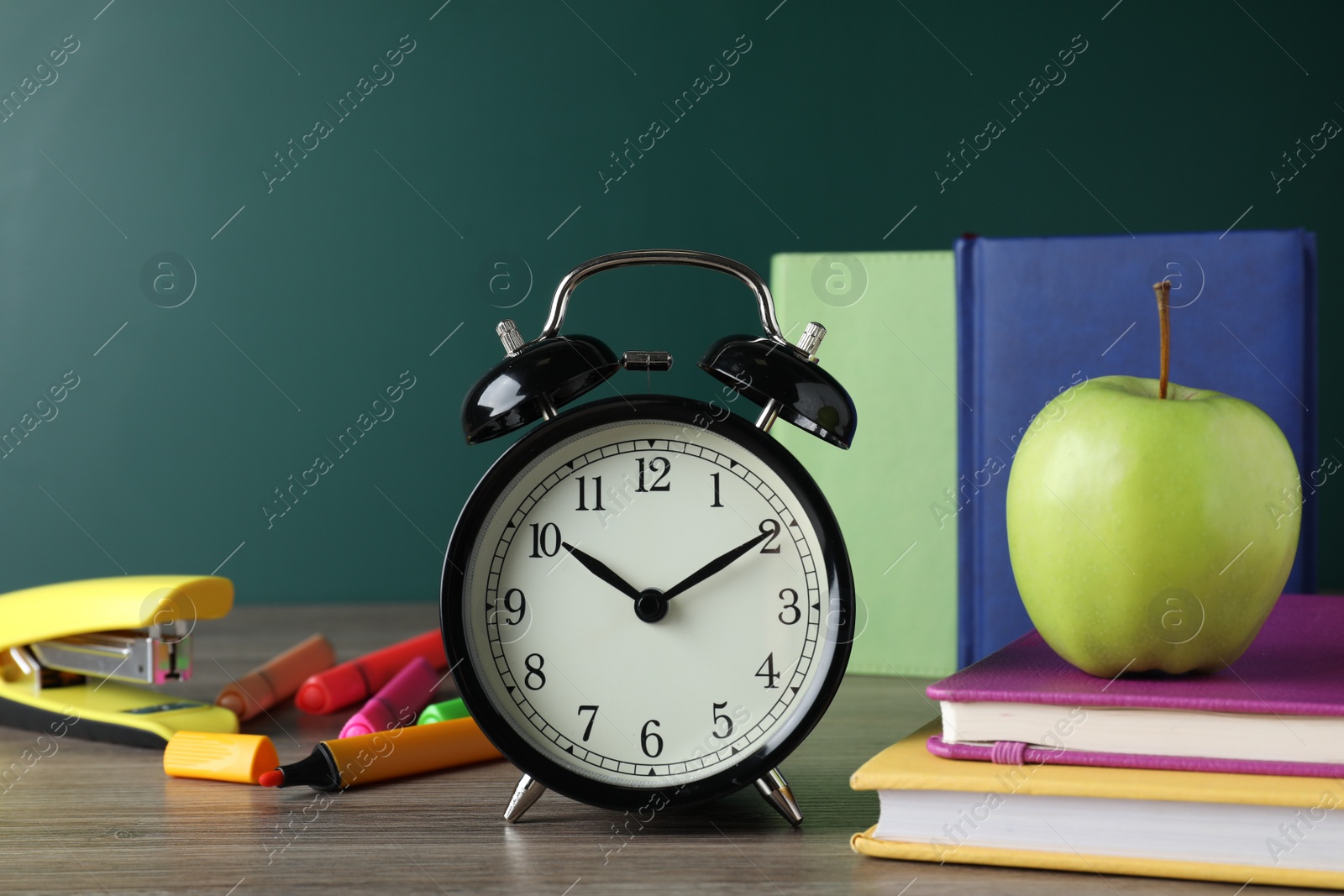 Photo of Alarm clock and different stationery on wooden table near green chalkboard. School time