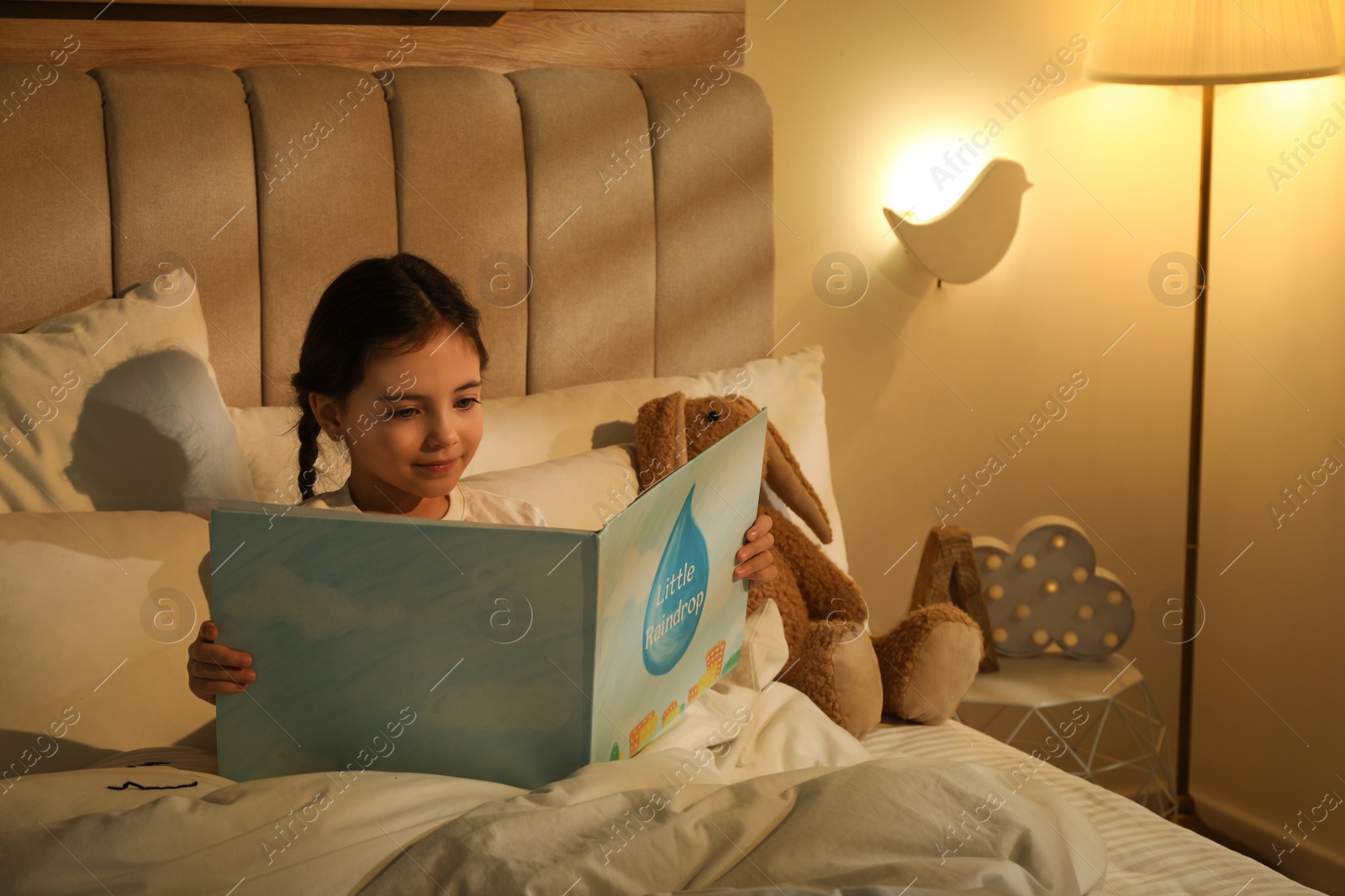 Photo of Little girl reading book in bedroom lit by night lamp