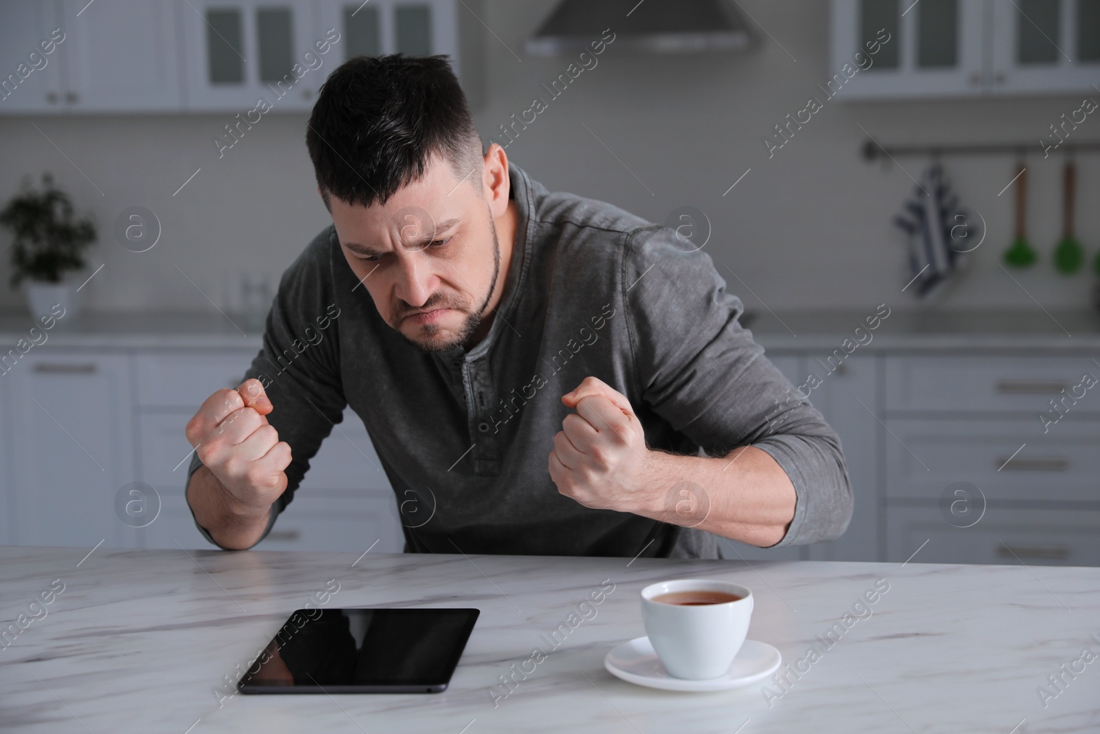 Photo of Emotional man with tablet at table in kitchen. Online hate concept