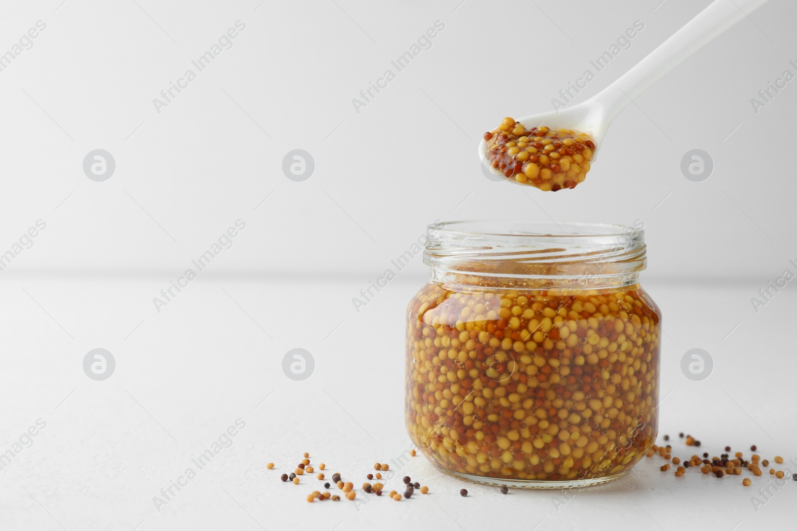 Photo of Taking whole grain mustard with spoon from jar on white table. Space for text