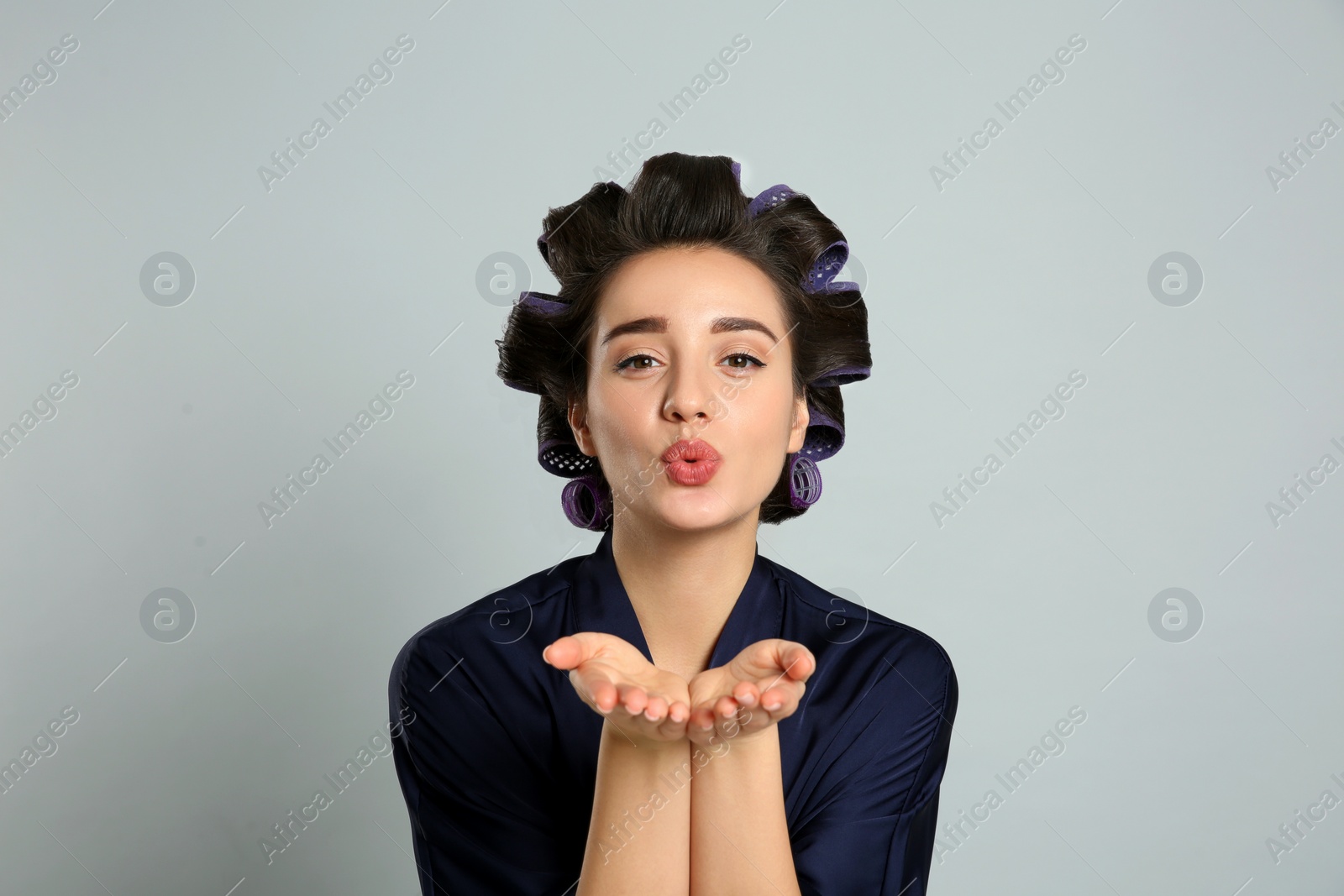 Photo of Young woman in silk bathrobe with hair curlers blowing kiss on grey background