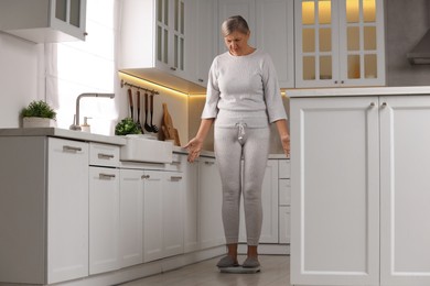 Menopause, weight gain. Concerned woman standing on floor scales in kitchen
