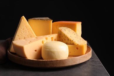 Photo of Wooden plate with different sorts of cheese on grey table against black background