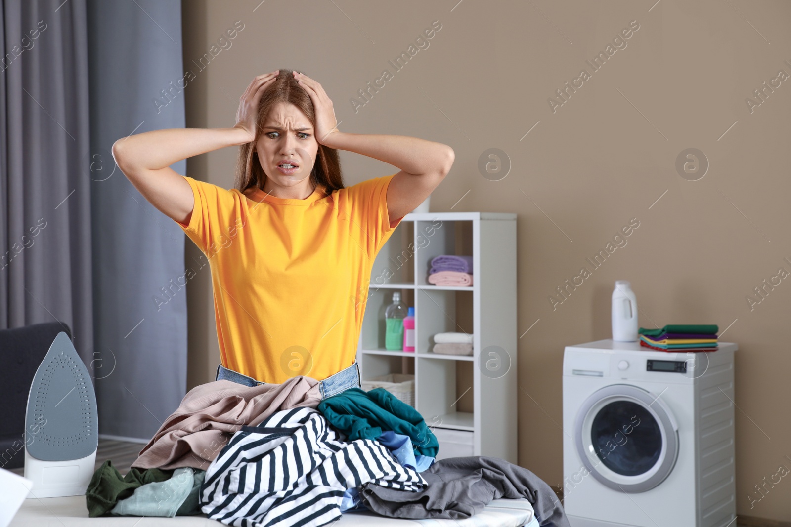 Photo of Emotional woman near board with iron and pile of clothes in bathroom. Space for text