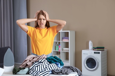 Emotional woman near board with iron and pile of clothes in bathroom. Space for text