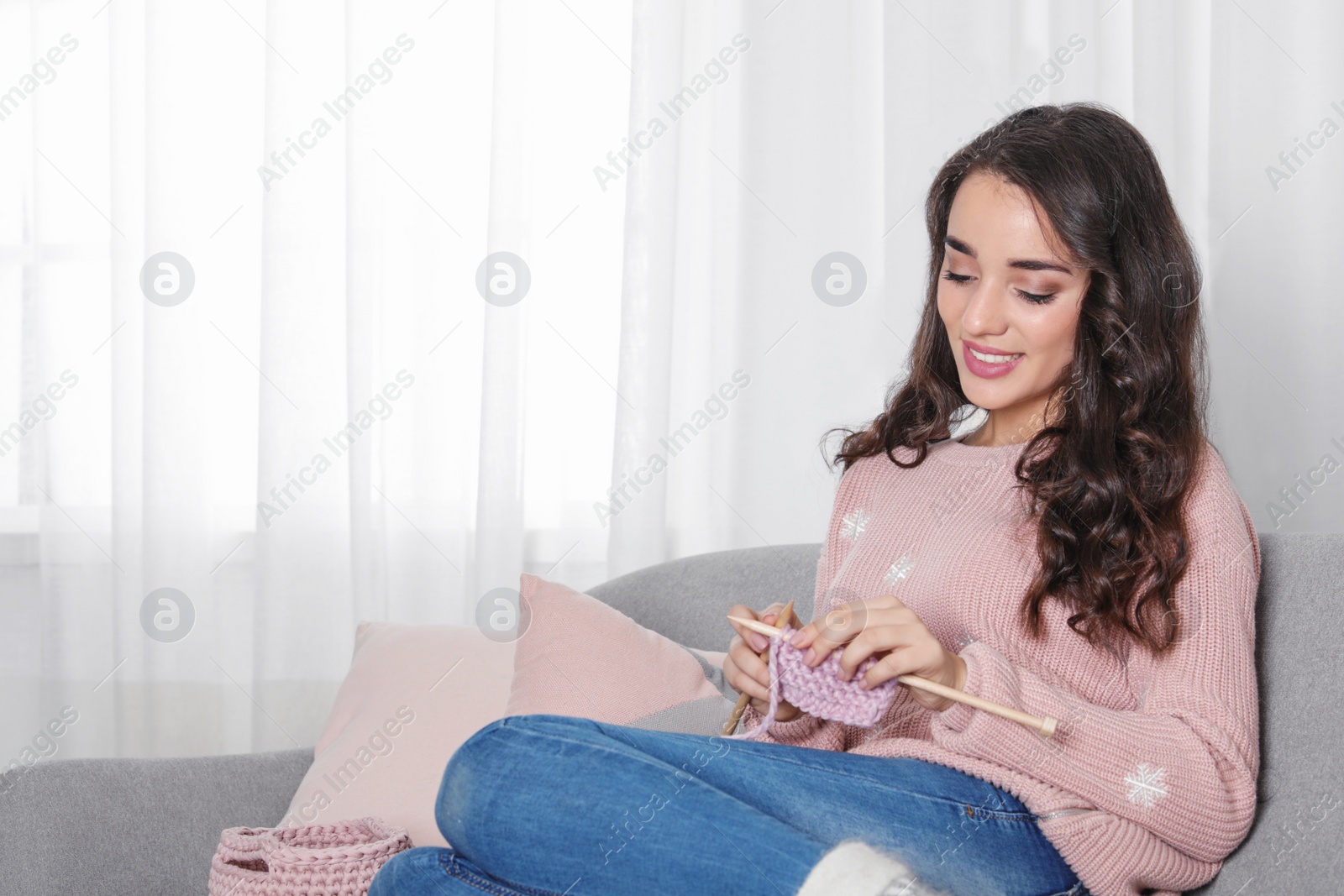Photo of Young woman in warm sweater knitting on sofa at home. Space for text