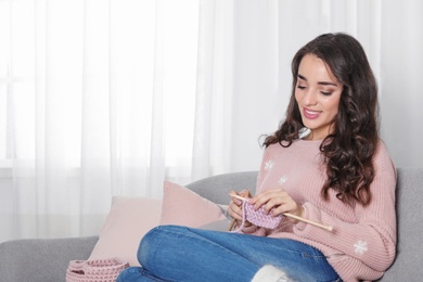 Young woman in warm sweater knitting on sofa at home. Space for text