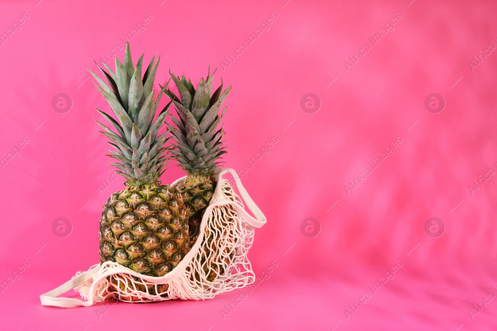 Photo of Whole ripe pineapples and net bag on pink background, space for text
