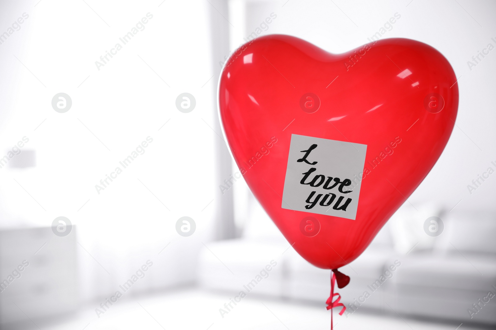 Photo of Red heart shaped air balloon with sticky note saying I Love You in room. Space for text