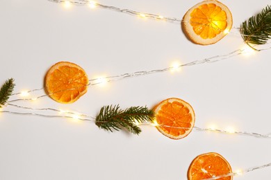 Photo of Christmas decor made of dry orange slices, festive lights and fir tree branches on white wall, closeup
