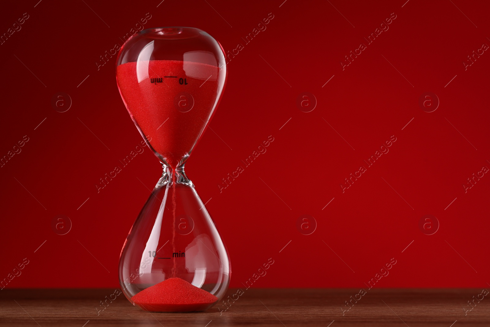 Photo of Hourglass with flowing sand on wooden table against red background, space for text