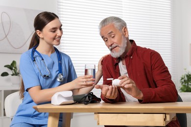 Photo of Young healthcare worker giving glass of water to senior man with pills at wooden table indoors