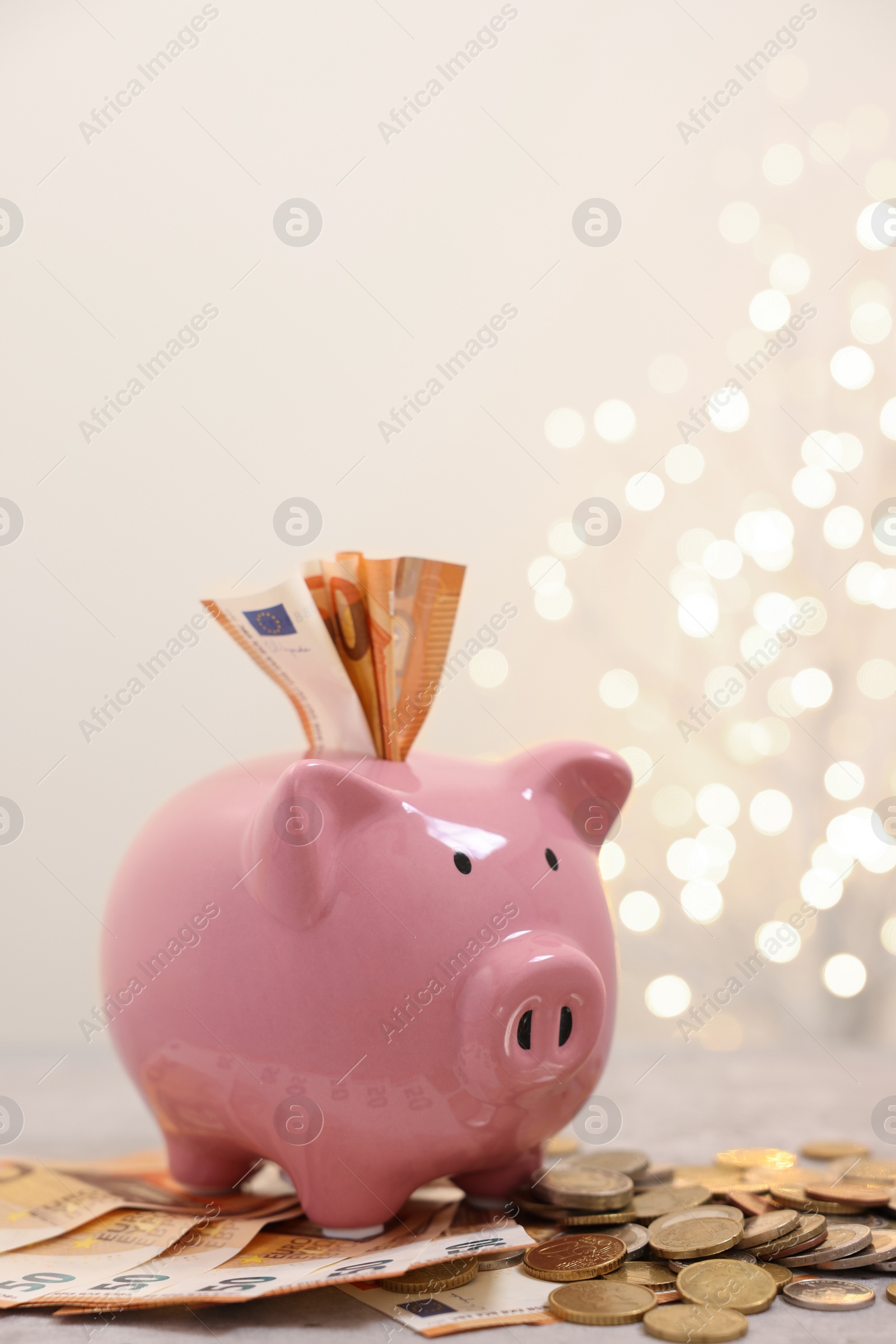 Photo of Piggy bank with euro banknotes and coins on grey table against blurred lights, space for text