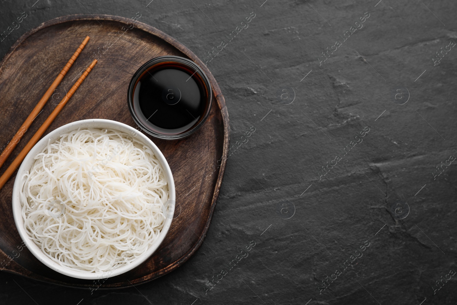 Photo of Bowl with cooked rice noodles, soy sauce and chopsticks on black table, top view. Space for text
