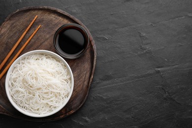 Bowl with cooked rice noodles, soy sauce and chopsticks on black table, top view. Space for text