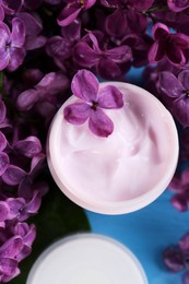 Photo of Jar of cream and lilac flowers on light blue table, flat lay