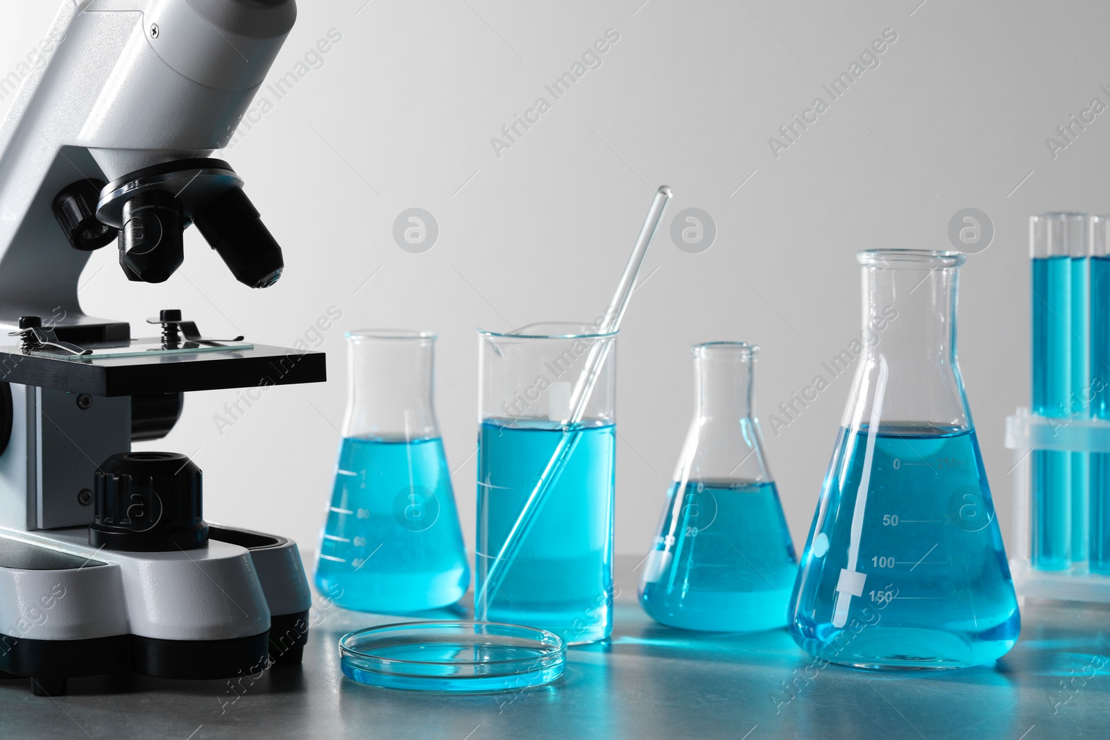 Photo of Different laboratory glassware with light blue liquid and microscope on table