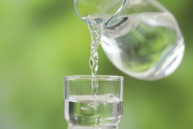 Pouring fresh water from jug into glass against blurred green background, closeup