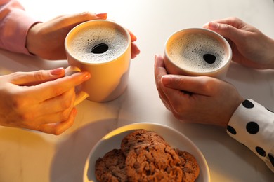 Photo of Women with cups of hot coffee and cookies at white marble table, closeup