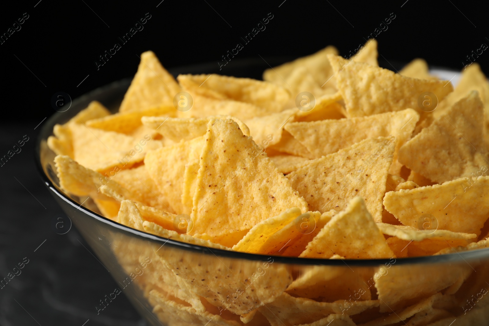 Photo of Glass bowl with tortilla chips (nachos) on black table, closeup