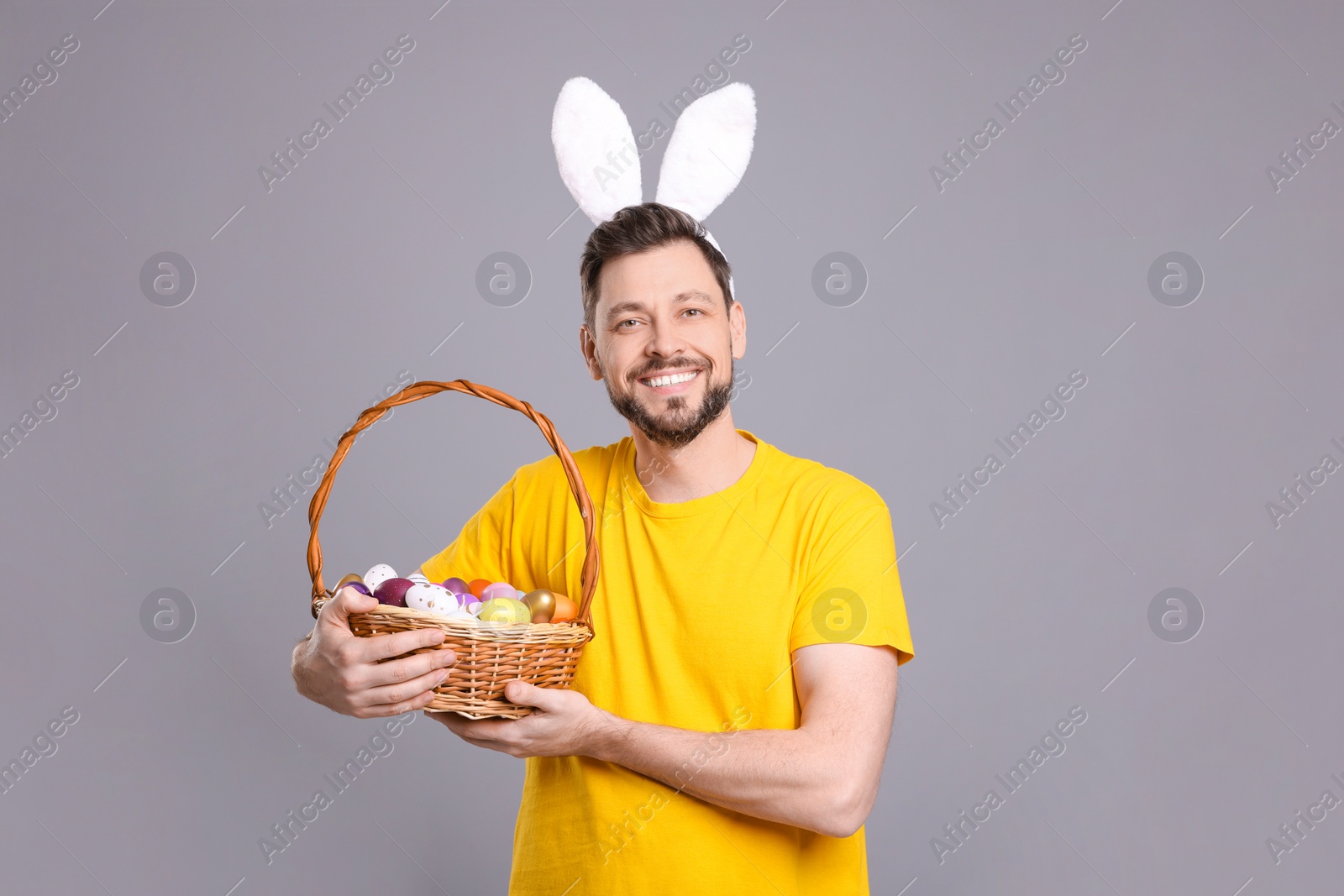 Photo of Happy man in bunny ears headband holding wicker basket with painted Easter eggs on grey background