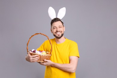 Photo of Happy man in bunny ears headband holding wicker basket with painted Easter eggs on grey background