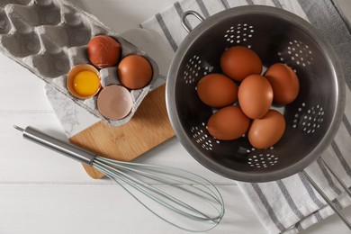 Photo of Fresh raw chicken eggs with colander, napkin and whisk on white wooden table, flat lay