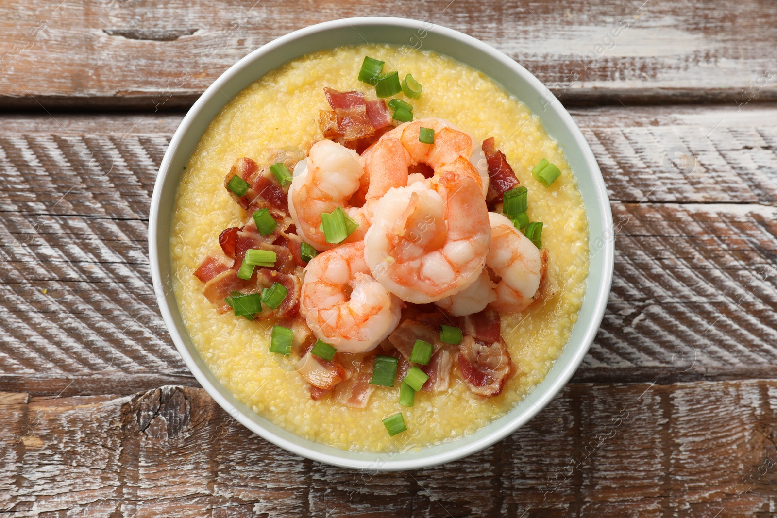 Photo of Fresh tasty shrimps, bacon, grits and green onion in bowl on wooden table, top view