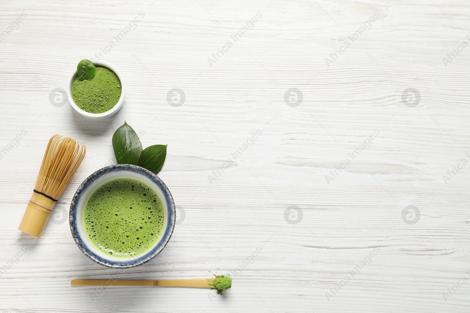 Photo of Cup of fresh matcha tea, bamboo whisk, spoon and green powder on white wooden table, flat lay. Space for text