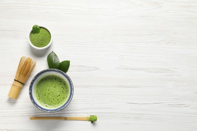 Photo of Cup of fresh matcha tea, bamboo whisk, spoon and green powder on white wooden table, flat lay. Space for text