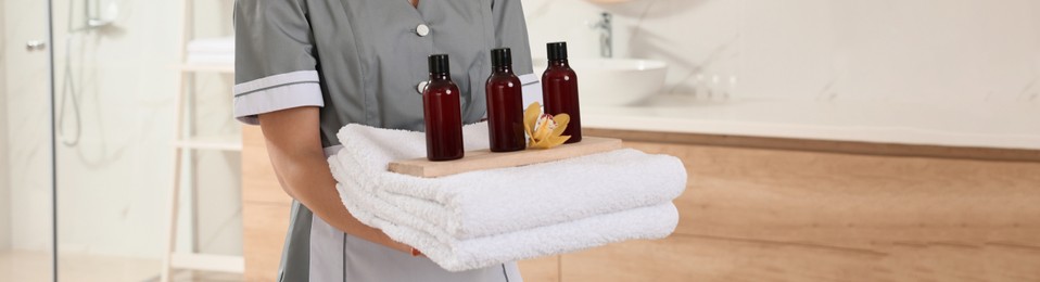 Chambermaid holding fresh towels with flower and shampoo bottles in hotel bathroom, closeup. Banner design
