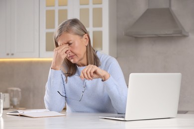Menopause. Woman with glasses suffering from headache at table in kitchen