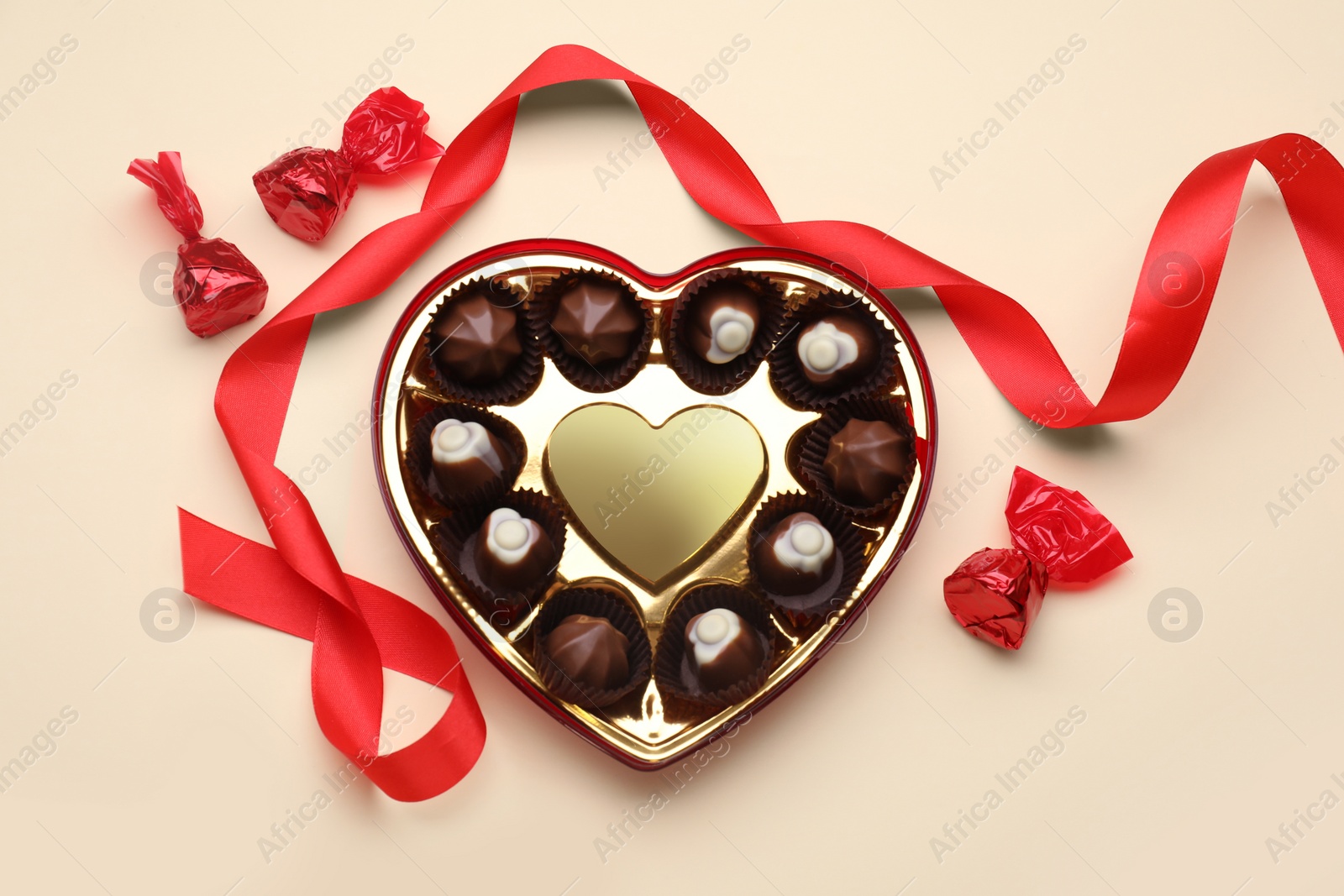Photo of Heart shaped box with delicious chocolate candies and ribbon on beige background, flat lay
