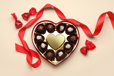 Heart shaped box with delicious chocolate candies and ribbon on beige background, flat lay