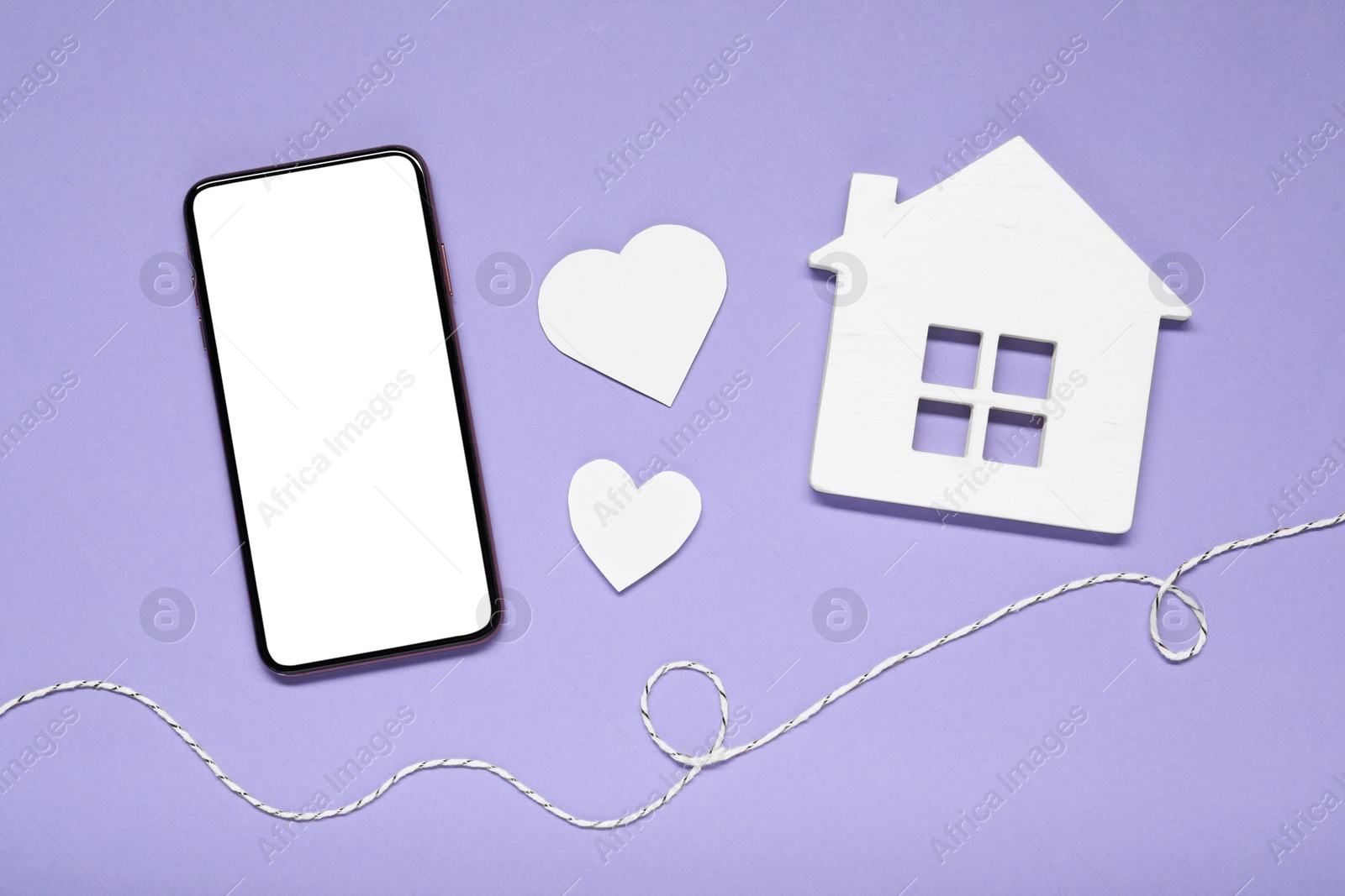 Photo of Long-distance relationship concept. Smartphone, white house, paper hearts and decorative cord on violet background, flat lay