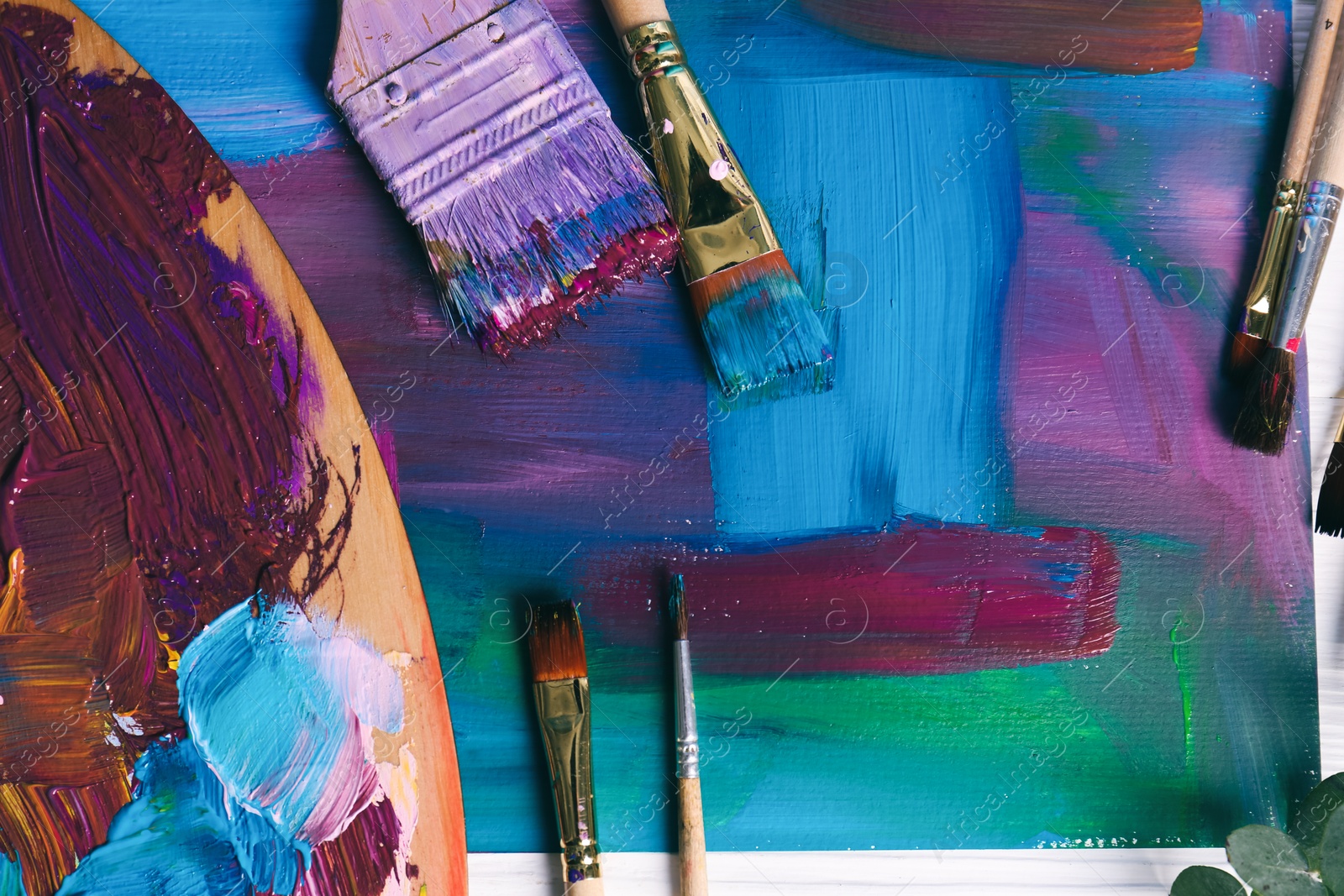 Photo of Canvas with colorful abstract painting, different brushes and wooden artist's palette on table, flat lay