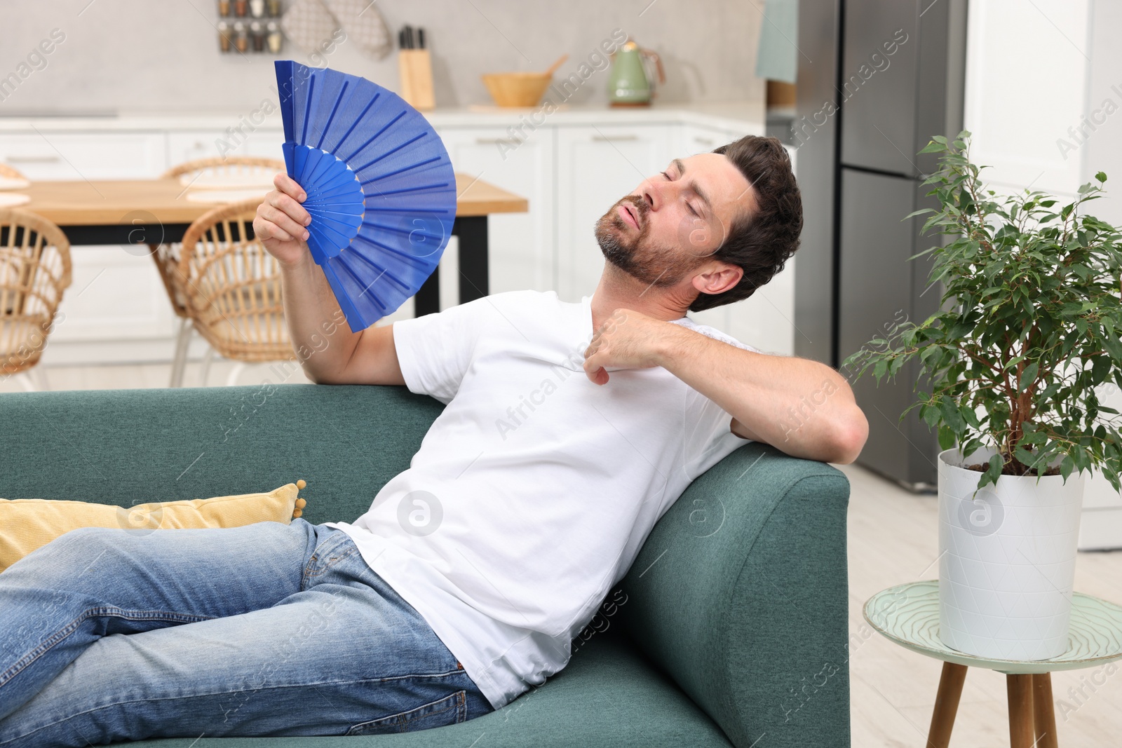 Photo of Bearded man waving blue hand fan to cool himself on sofa at home