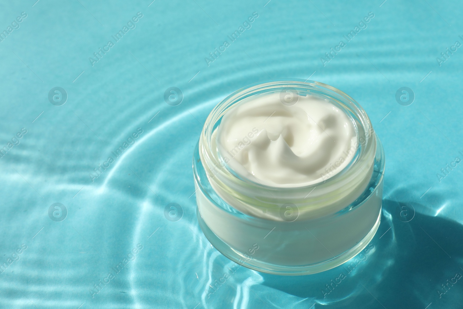 Photo of Jar with moisturizing cream in water on light blue background, closeup. Space for text