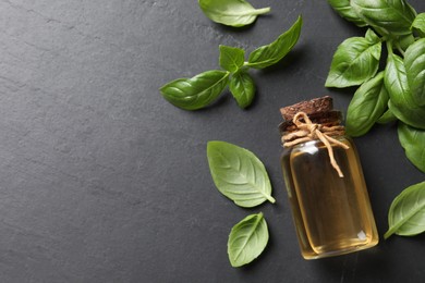 Photo of Bottle of essential basil oil and fresh leaves on dark grey table, flat lay. Space for text