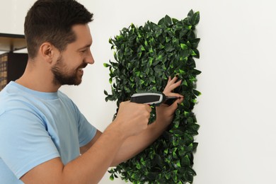 Man with screwdriver installing green artificial plant panel on white wall in room