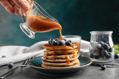 Woman pouring caramel syrup onto fresh pancakes with blueberries at grey table, closeup